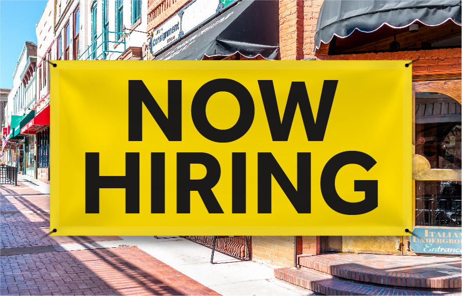 Yellow banner with black text that says "NOW HIRING" in front of a street corner with businesses in the background